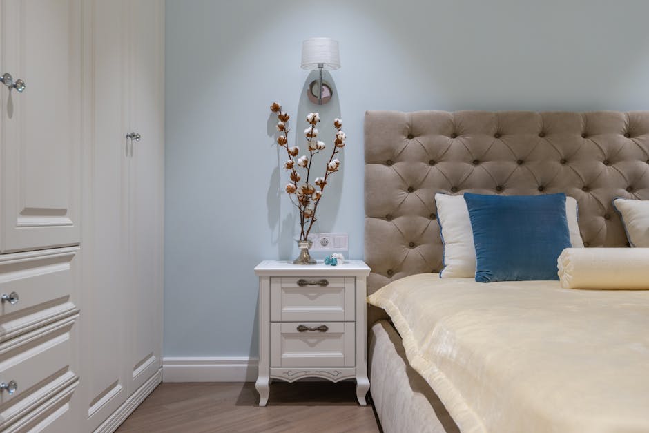 White classic styled wardrobe and nightstand decorated with vase with cotton plants placed near comfortable bed with cushions in cozy apartment