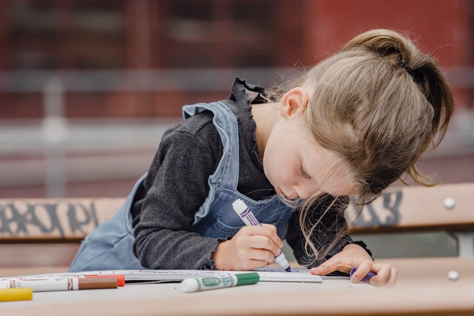 Adorable preschool girl in casual clothes sitting at table on schoolyard and holding marker while drawing illustration