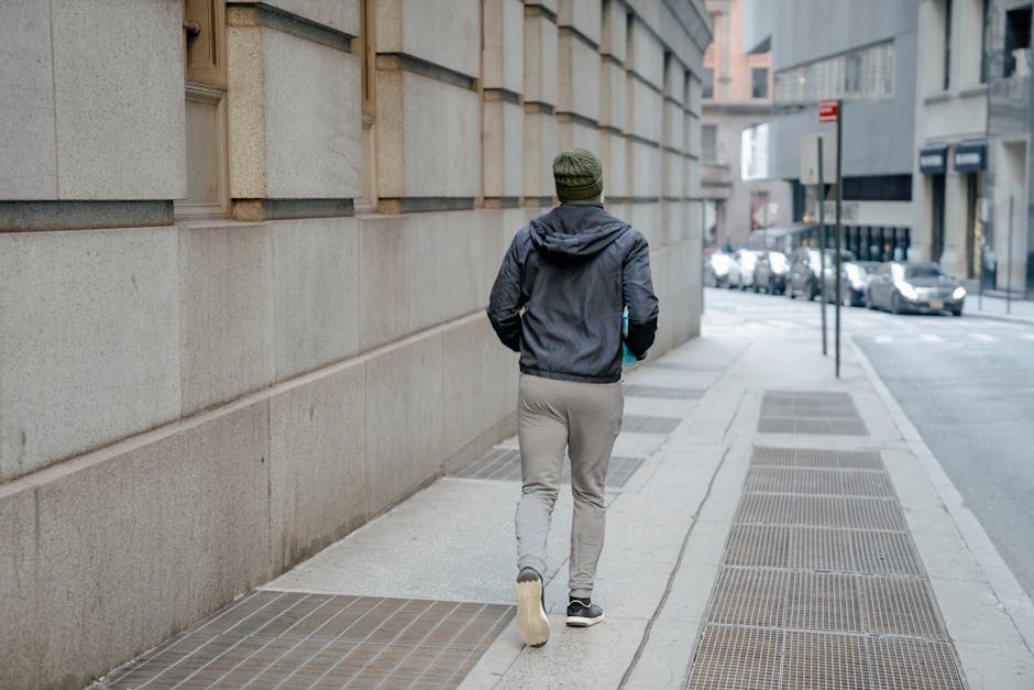 Back view of full body anonymous male wearing windbreaker jacket and gray sweatpants and knitted hat runnning on paved walkway along building