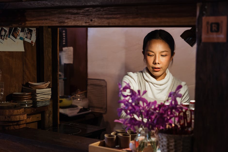 Content young ethnic female worker wearing apron standing behind counter with flower bouquet and looking down while working in cafe with rustic wooden interior in dark light in evening