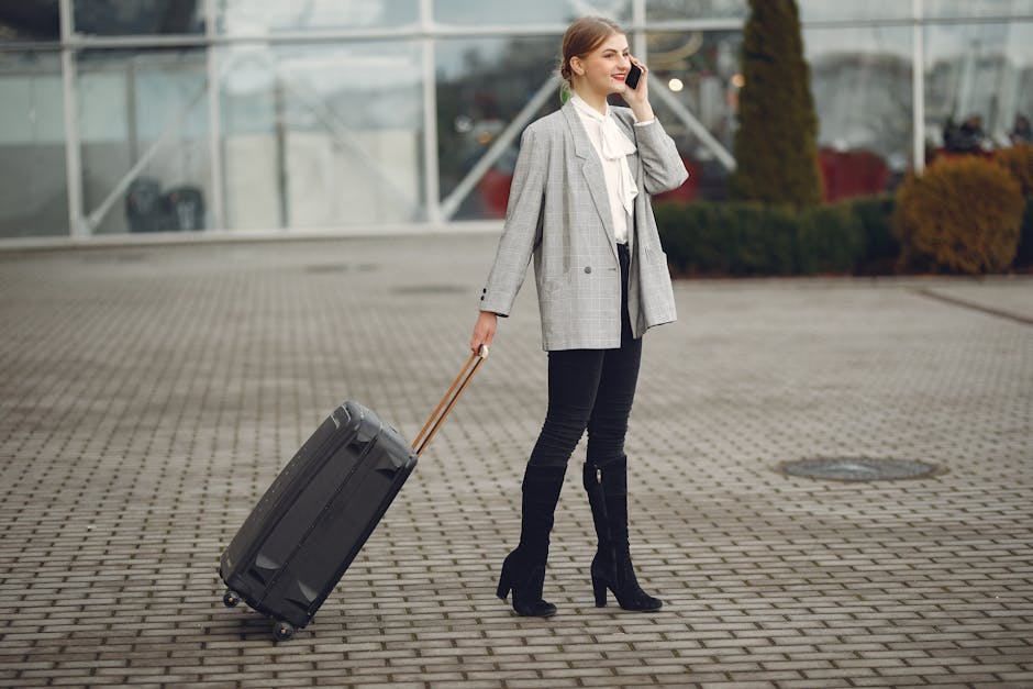 Full body of elegant female tourist in stylish clothes standing with suitcase and calling taxi by phone after arrival in modern city