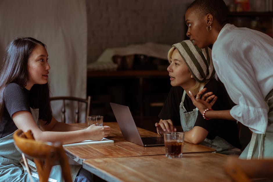 Serious multiethnic waitresses in aprons sitting at wooden table with modern netbook and notebook while discussing business project