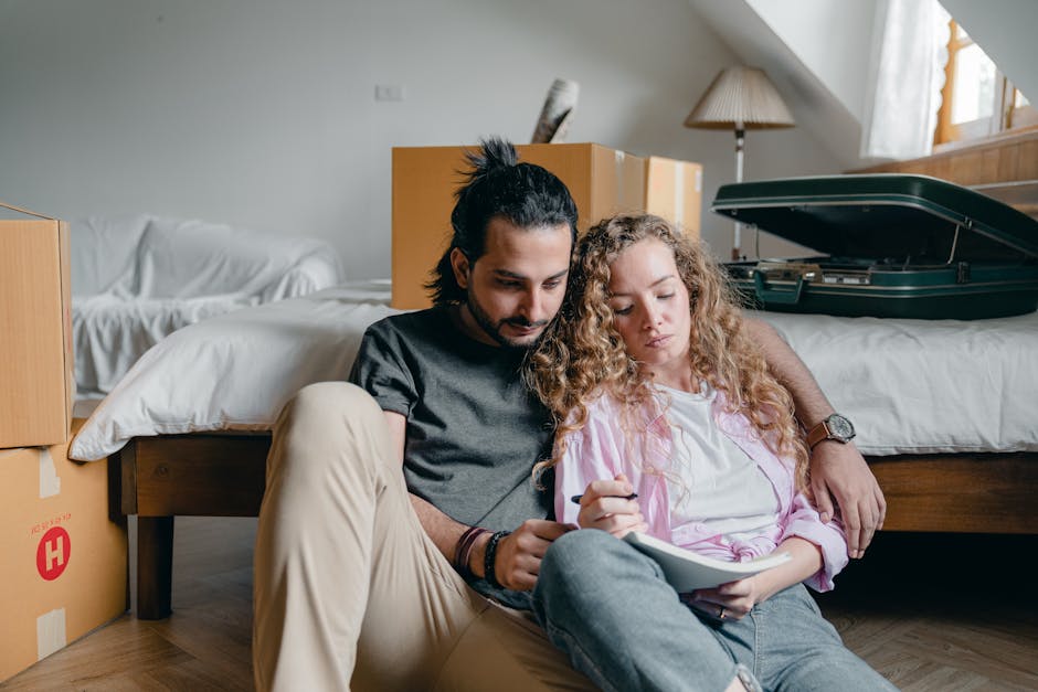 Thoughtful male and female in casual wear sitting near bed among boxes together and taking notes while moving house