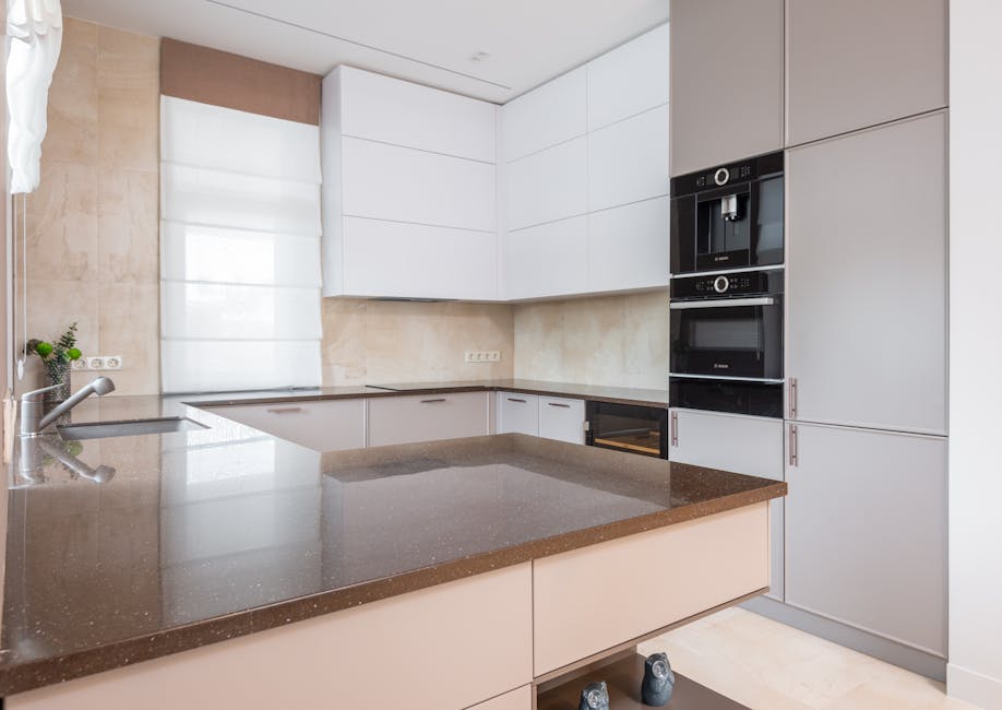 Contemporary kitchen with white and beige cupboards and built in appliances and cutting table laid with stone countertop and metal sink