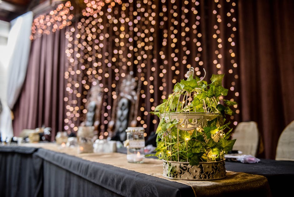 Long banquet table under dark tablecloth with creative green plant and candles located near closed curtains with shining garlands