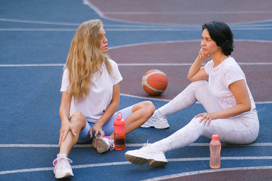 Full length of confident young ladies in sportswear speaking while having break on court after exercising with water bottles near basketball in daylight