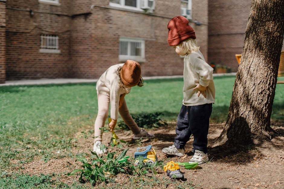 Unrecognizable little kids in casual outfits and brown hats playing with toys on backyard beside big tree