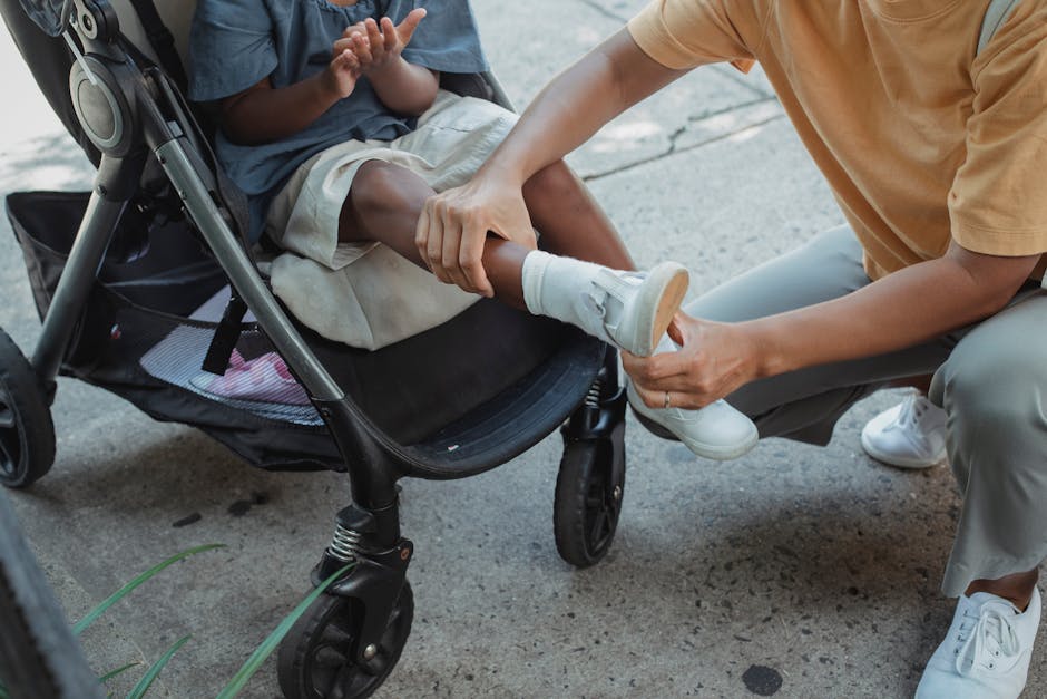 From above of crop anonymous ethnic caring mother putting shoes on little daughter in baby carriage on asphalt pavement