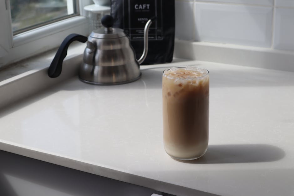 High angle glass of delicious iced latte and metal coffee kettle placed on white table in kitchen