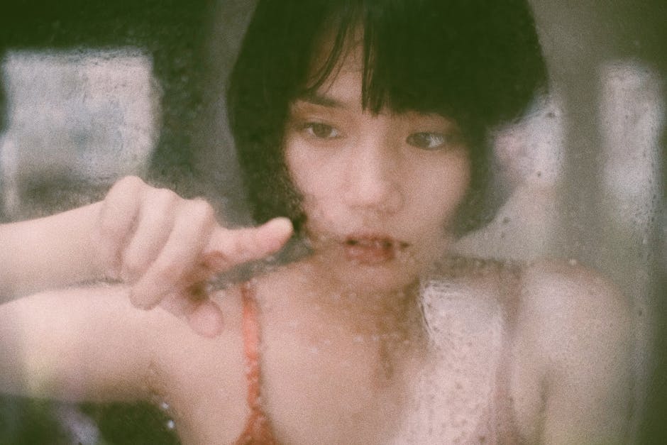 Through window of young Asian woman drawing on  wet glass with finger in soft light