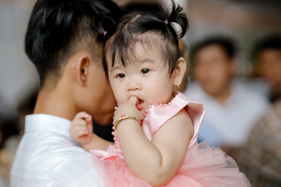 Adorable Asian toddler with ponytails in pink dress sitting on hands and sucking finger