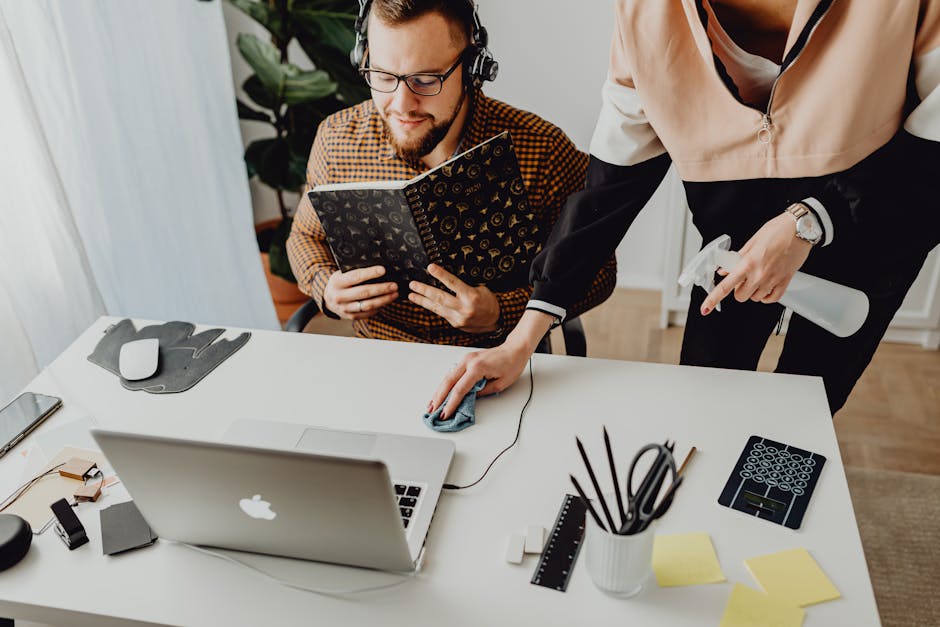 Woman Cleaning the Desk While Man Sits and Uses Laptop