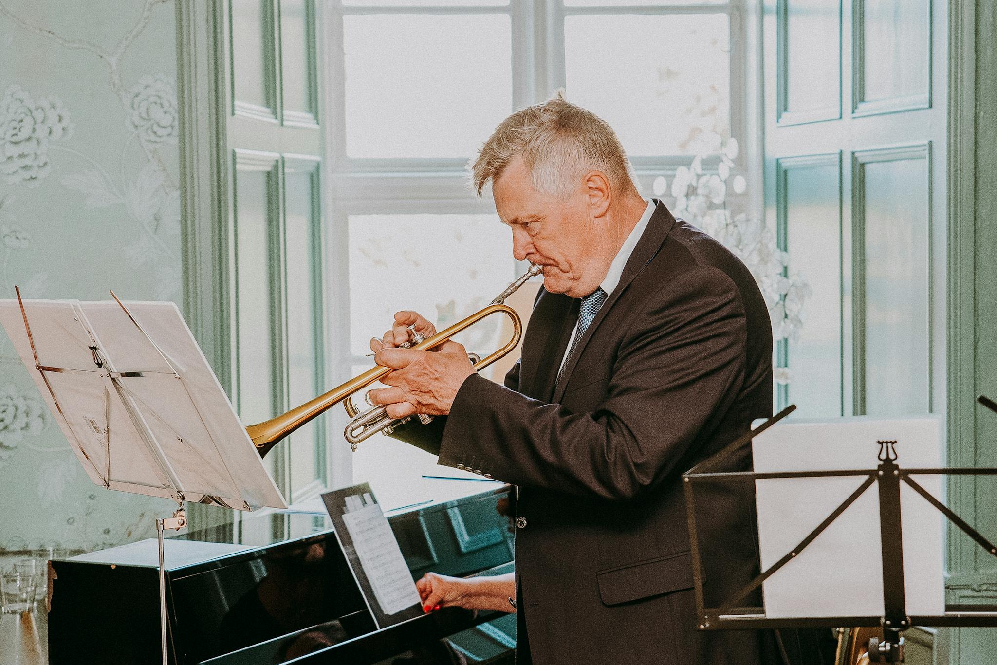 Side view of concentrated senior musician with grey hair in dark elegant suit looking at notes on sheet and playing trumpet next to pianist