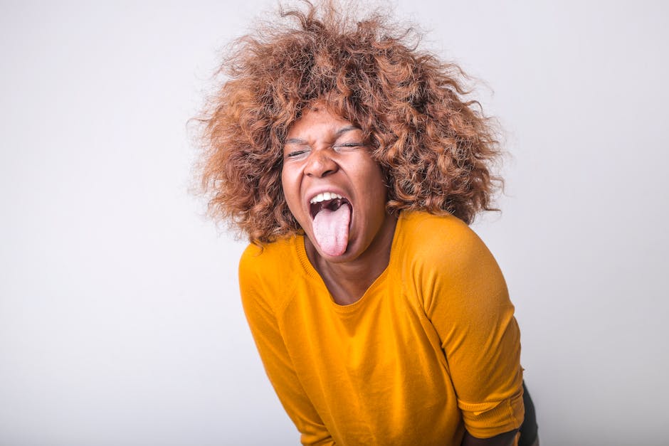 Red haired African American female in vivid yellow sweater standing with eyes closed and tongue out against gray background in modern studio