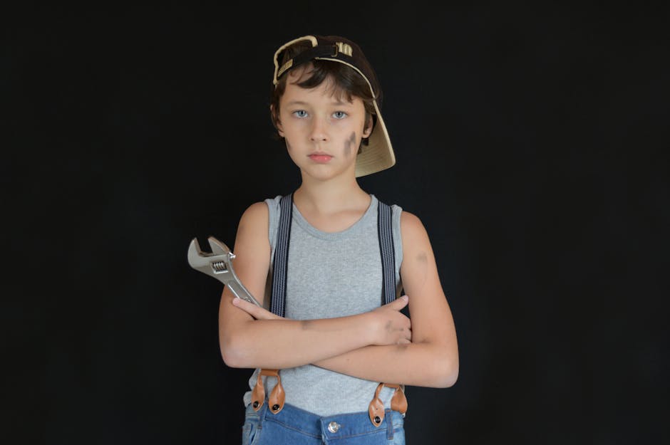 Serious preteen boy with dirty face wearing vehicle technician outfit standing with arms folded and wrench against black background while looking at camera