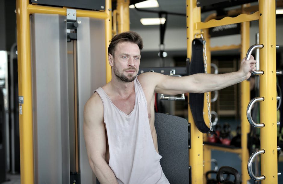 Serious bearded male athlete in casual sportswear sitting near modern exercise machine during break in workout in modern fitness center and looking away