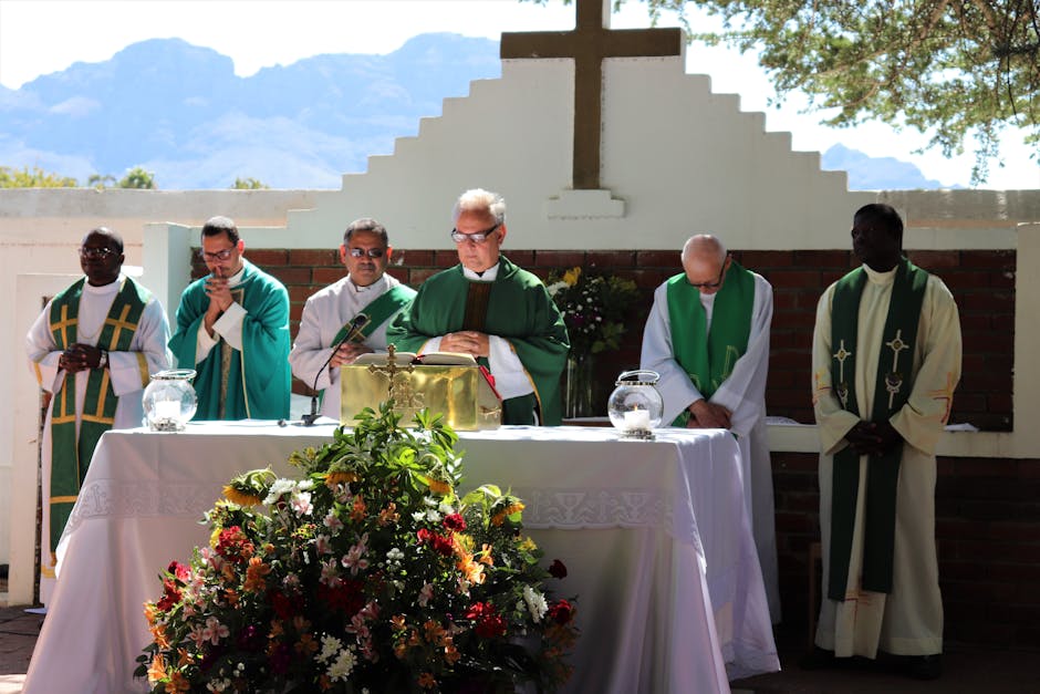Elderly priest in eyeglasses and catholic vestment standing with prayer hands near table with Bible and multiethnic deacons during liturgy process behind mountains in sunlight