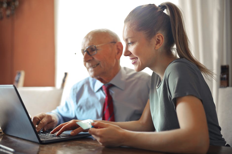 Young woman in casual clothes helping senior man in formal shirt with paying credit card in Internet using laptop while sitting at table