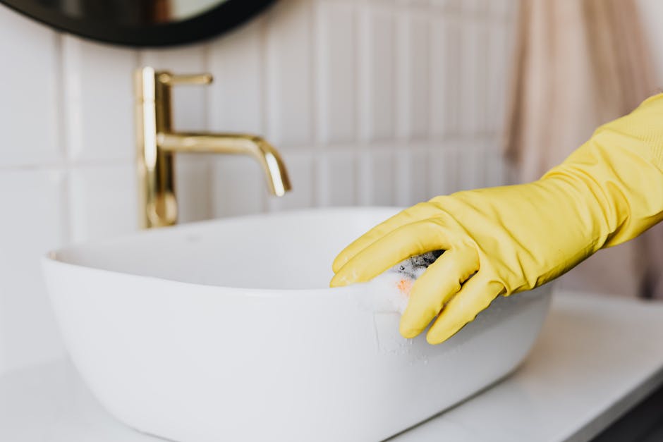 Crop faceless person in yellow latex protective glove cleaning surface of white stylish sink in contemporary bathroom interior