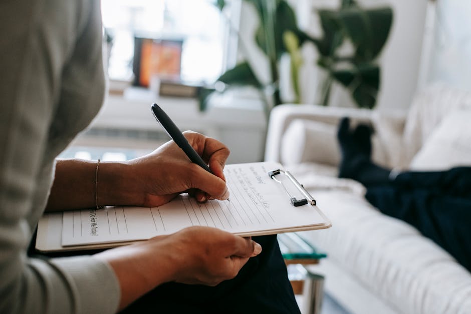 Unrecognizable ethnic female therapist taking notes on clipboard while filling out form during psychological appointment with anonymous client lying on blurred background