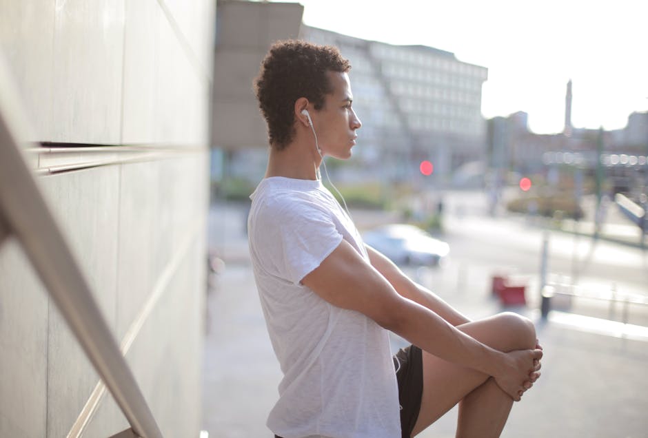 Side view of calm thoughtful African American male athlete in earphones and sportswear looking away and stretching leg while standing alone and listening to music on street near modern building in city