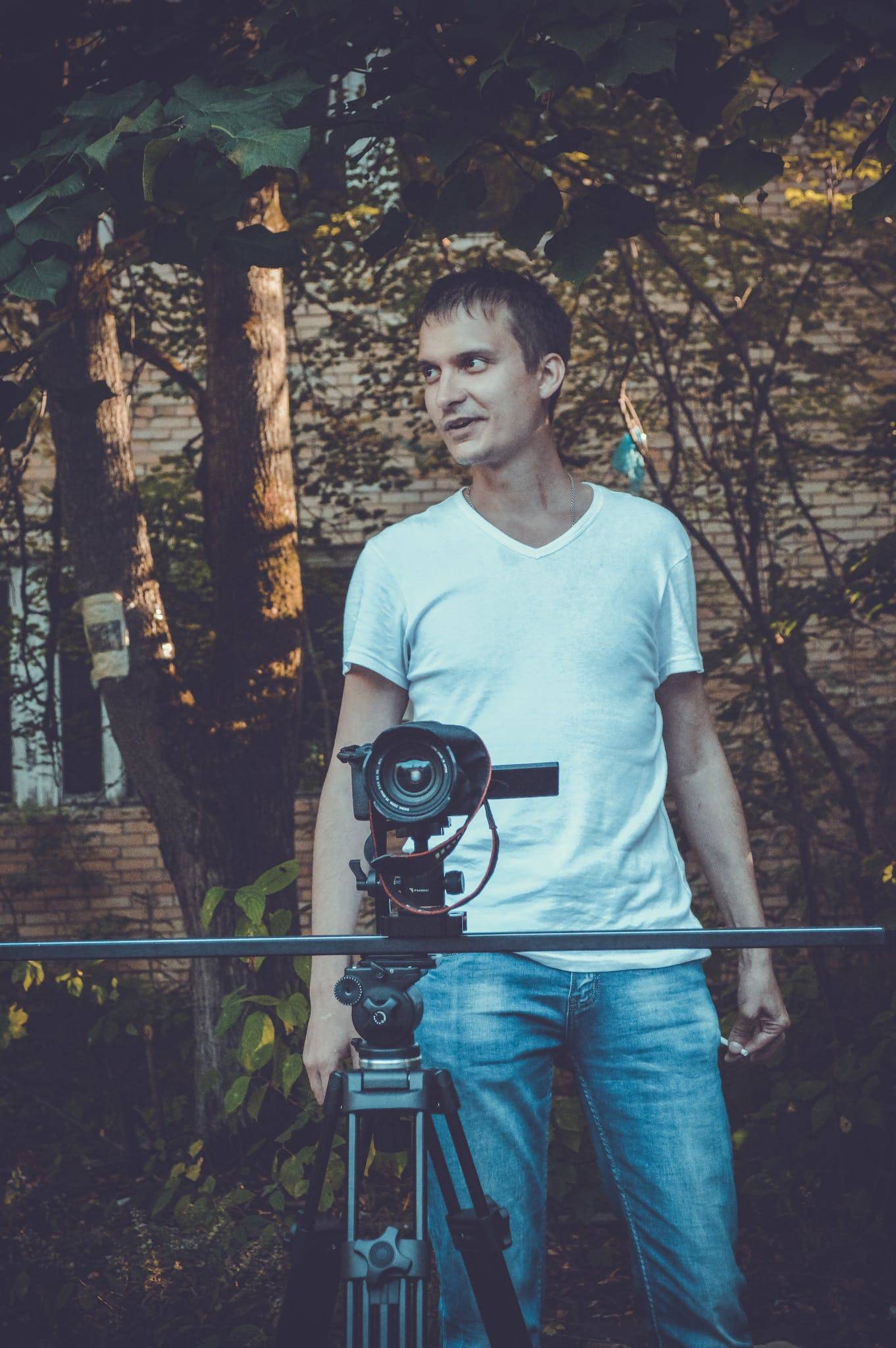 Camera man in white shirt and jeans looking away while taking video on professional equipment on tripod