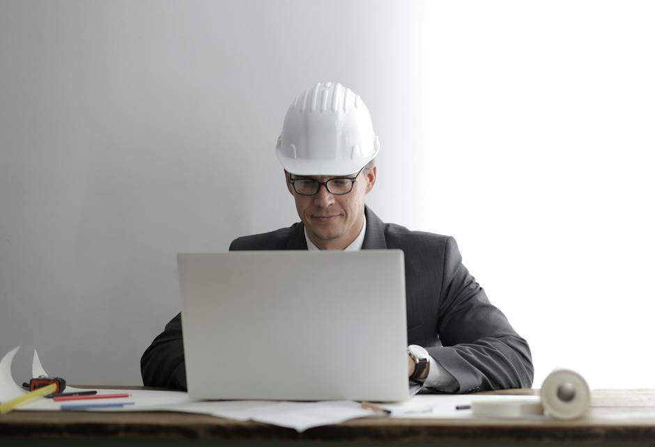 Serious concentrated engineer in hardhat and formally suit sitting at table with blueprints while working on laptop against gray background