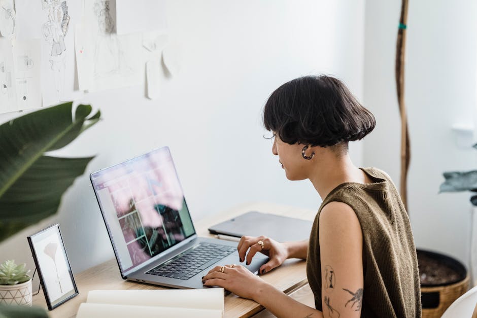 Side view of young female worker sitting at table and working on laptop in light studio