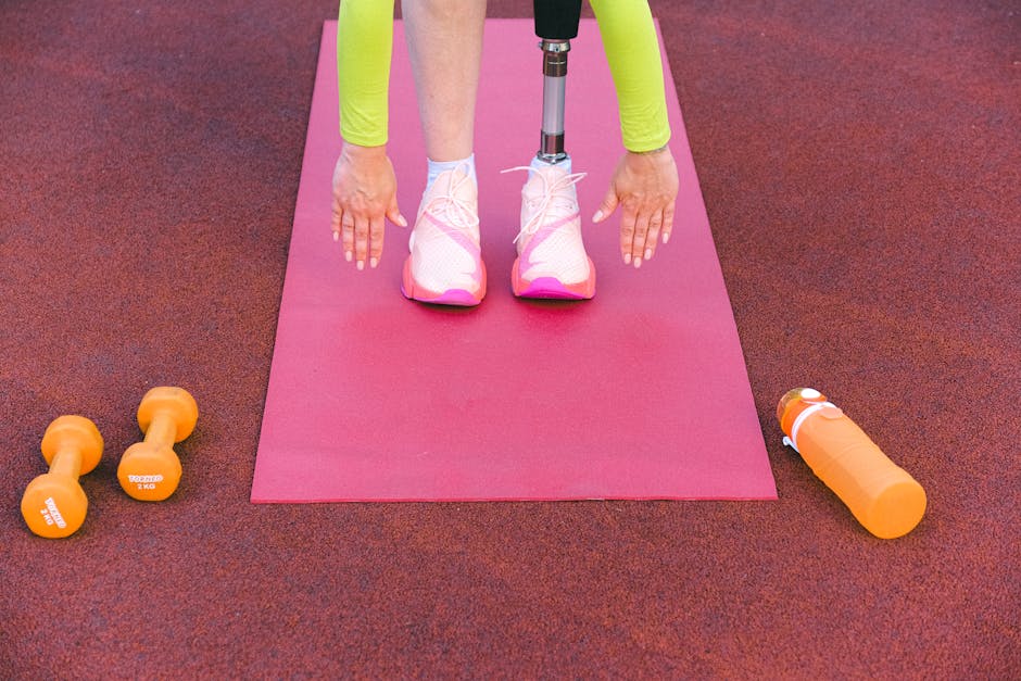 Unrecognizable female athlete with leg prosthesis bending forward and stretching while trying ti touch ground during training on sports ground with dumbbells