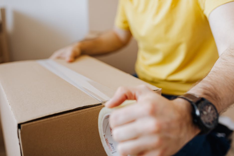 Crop anonymous male in casual wear using cling tape to seal cardboard container with belongings during relocation