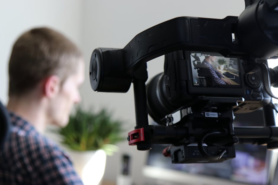 Black Video Camera Turn on Next to Man Wears Black and Gray Shirt