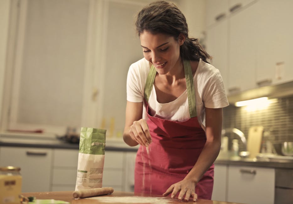 Low angle of positive young female in apron sprinkling flour over table while preparing food in modern light kitchen