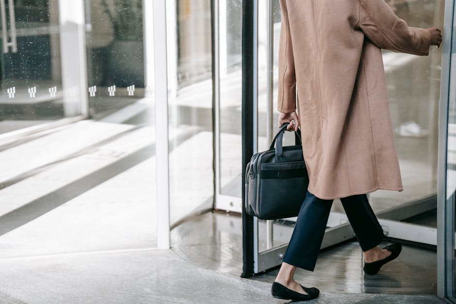Back view of crop stylish female manager in elegant clothes with briefcase in hand entering modern building through revolving door while going to work