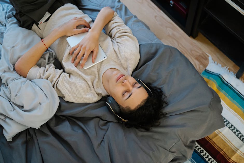 From above young Hispanic guy with tablet listening to music with closed eyes while chilling on comfortable bed at home