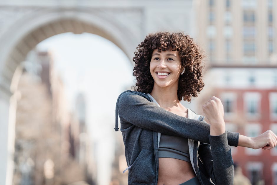 Happy ethnic sportswoman in activewear stretching arms and looking away while training on street with historic arch on blurred background