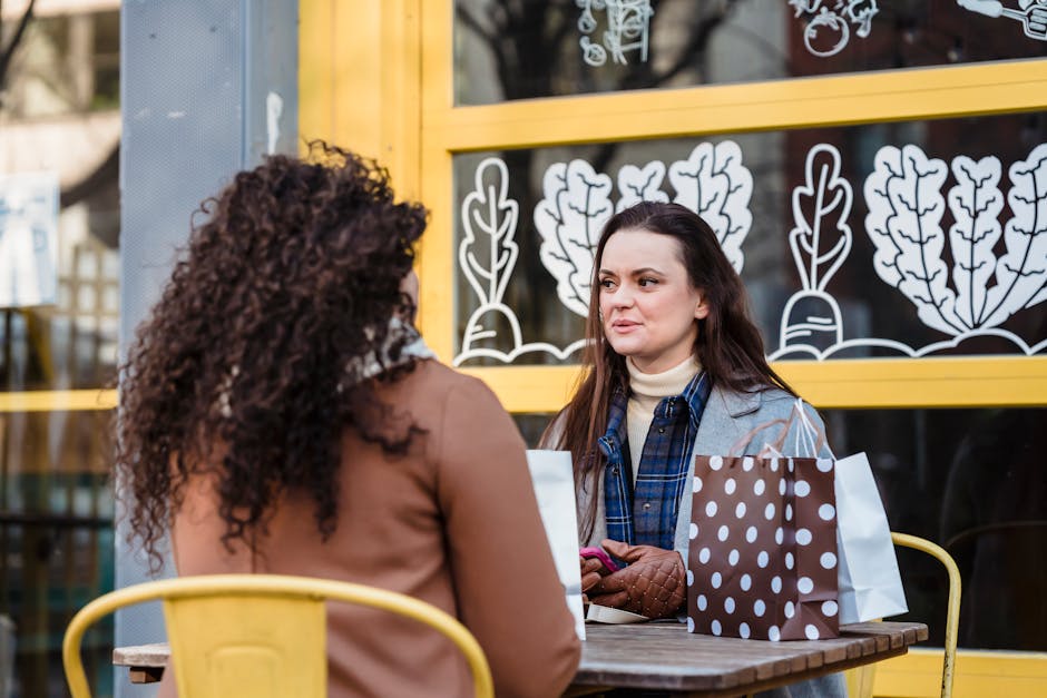 Young ladies wearing warm coats and gloves while sitting at table in chairs and talking in city street near building and shopping packages with smartphone in hand in daytime