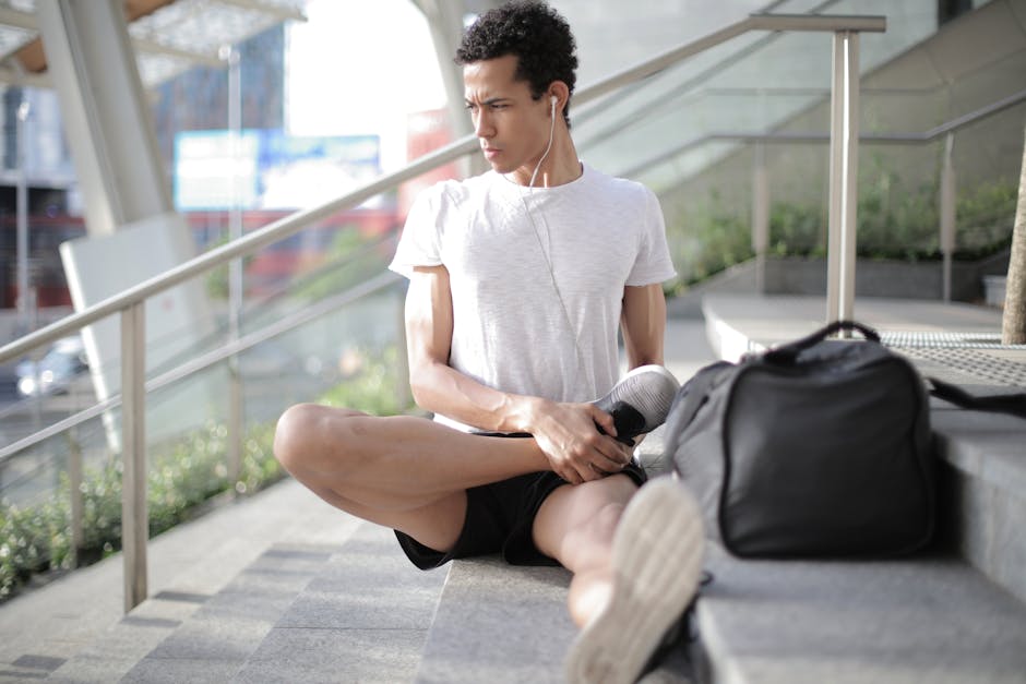 Young ethnic athlete in earbuds stretching body while sitting on stairs at entrance of contemporary building