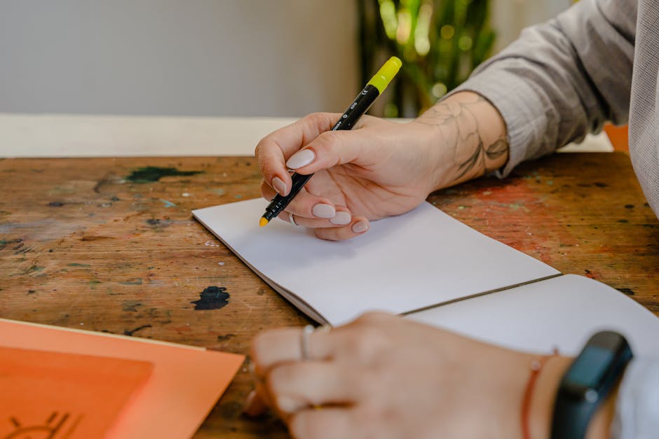 Crop unrecognizable designer in smart watch sitting with yellow marker and open empty album at wooden table with ink blots on surface