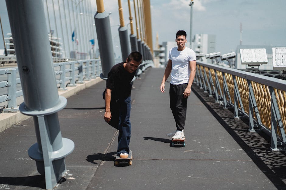 Full body of young ethnic men skateboarding on asphalt bridge with metal constructions under cloudy sky