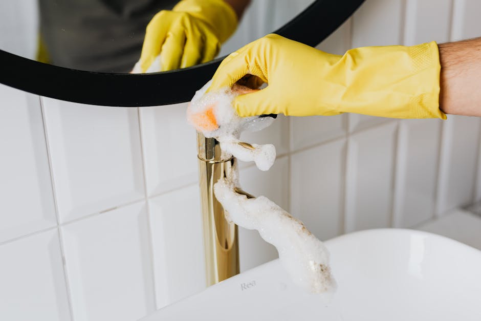 Anonymous person wearing yellow latex gloves washing golden faucet with sponge in modern bathroom while cleaning house