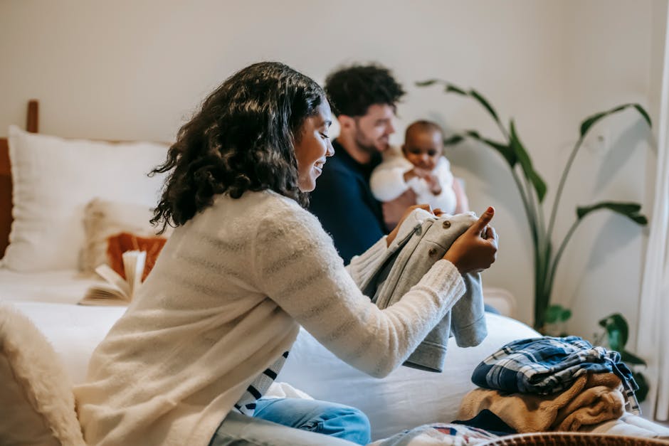 Positive African American mom organizing kid clothes sitting on bed nae farther cuddling cute baby in bedroom