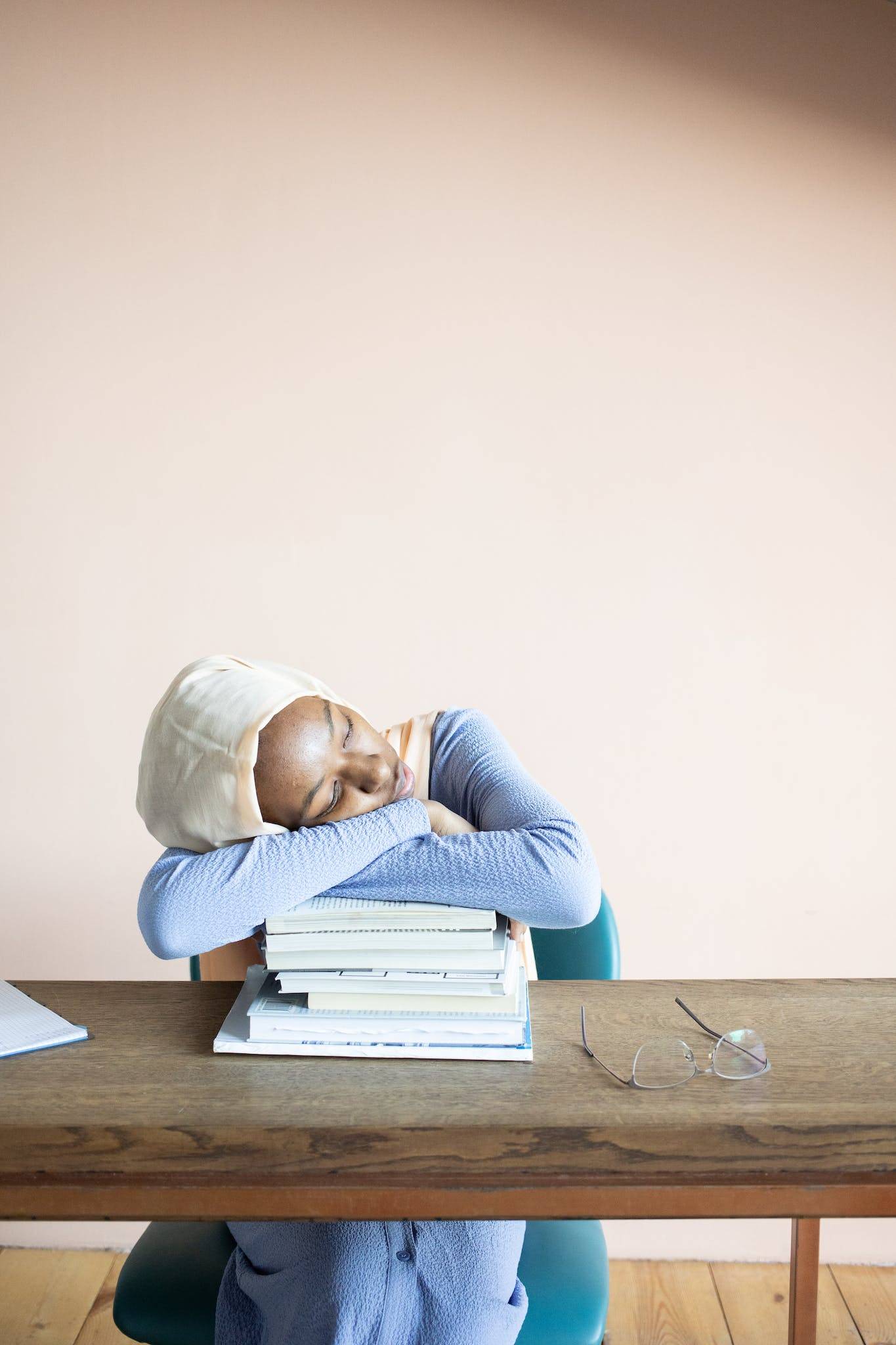 Exhausted African American female student in hijab having nap on stack of textbooks after studies