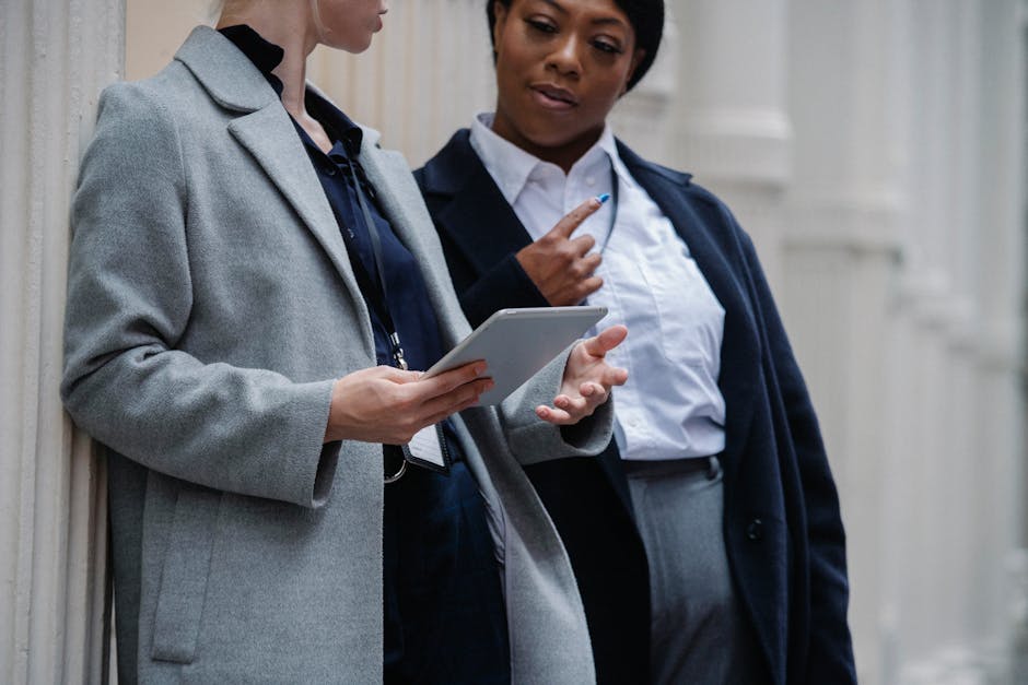 Crop smart multiethnic female colleagues in formal clothes and badges leaning on columns of aged building and sharing tablet during project discussion