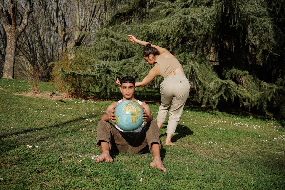 A Woman Dancing Behind a Man Sitting on Grass Holding a Globe
