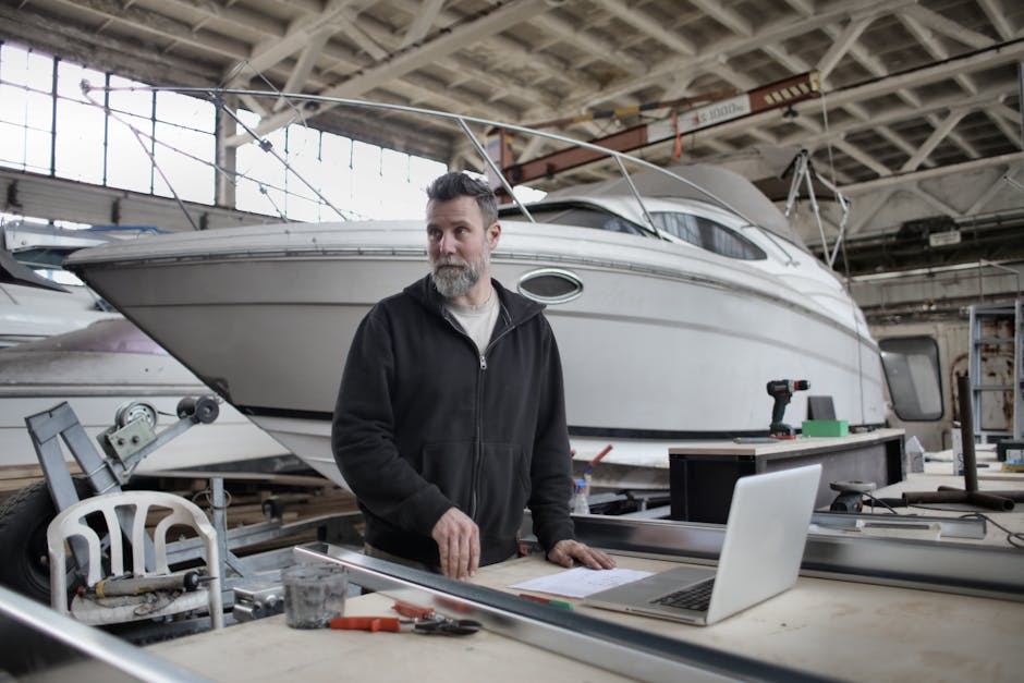 Adult bearded man in casual clothes using Mac while working with metal details near boat in workshop and looking away