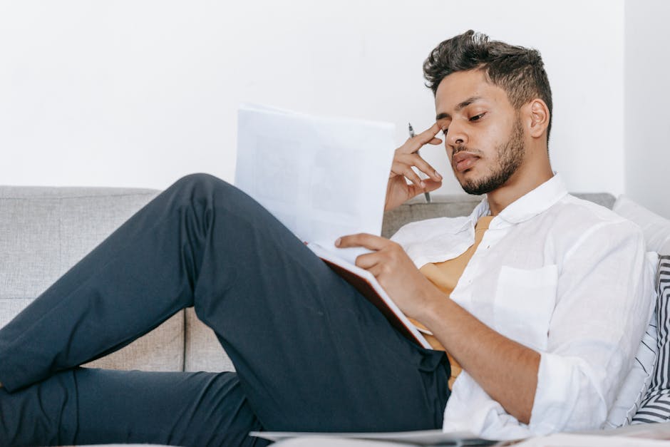 Pensive ethnic male worker touching temple while lying with agenda and papers on sofa in house room