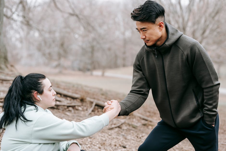 Young Asian male in sportswear giving hand to plump female fell on ground while training together in autumn leafless forest