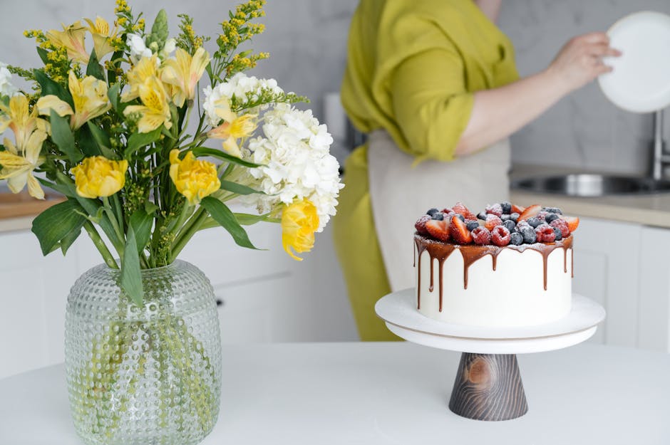Side view of crop unrecognizable woman in apron washing plates in kitchen standing near table with appetizing cake decorated with fresh assorted berries and bunch of flowers in vase