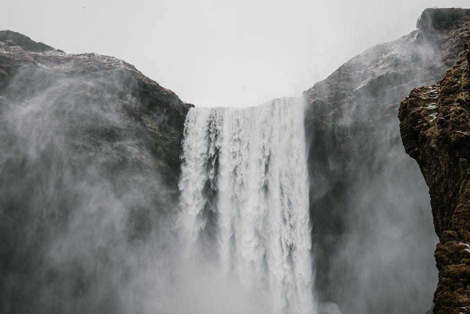 From below of majestic landscape with waterfall flowing through cliffs and water spray in air under gray sky during cold day