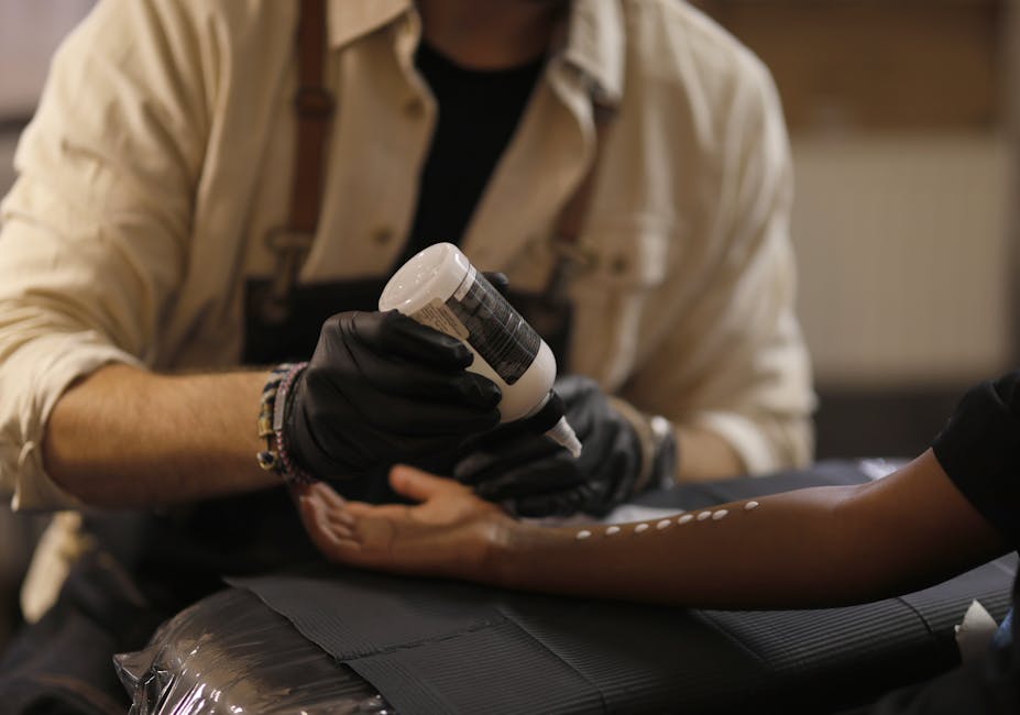 Unrecognizable crop male tattooist in black latex gloves and apron pouring tattoo transfer gel on arm of black female client in modern salon